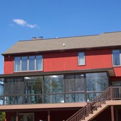 J & D Gutters - Large elevated screened-in porch attached to a red two-story home, featuring dark framing, expansive mesh panels, and a slope...