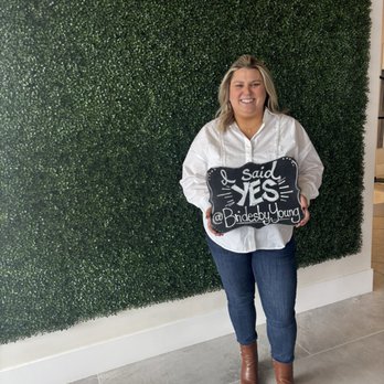 Brides By Young - A woman with blonde hair, wearing a white shirt, jeans, and brown boots, stands in front of a green hedge wall with the sign '@BridesbyYoung' and 'Yes' written on it. - Bridal Near Me - Schaumburg, Illinois