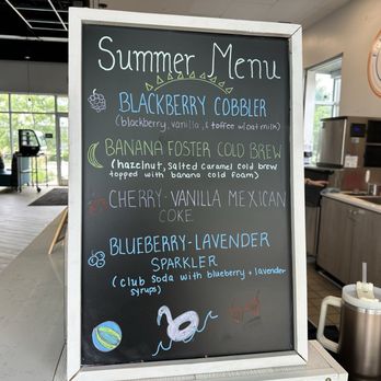 Epic Coffee - A chalkboard menu titled 'Summer Menu' lists various summer drinks at a coffee shop, including blueberry cobbler, banana foster cold brew, cherry-vanilla Mexican coke, and blueberry-lavender sparkler. The menu features colorful handwritten text and small illustrations, with a background showing part of the coffee shop interior, including seating areas and a counter. - Juice Bars & Smoothies Near Me - Carmel, Indiana
