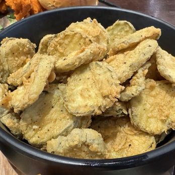 Main Event Tempe - Fried pickles! - A black bowl contains fried pickle slices, which are golden and crispy. To the side, there is a small container of creamy dipping sauce, and a glass with a beverage that appears to have lemon or orange slices. The setting suggests a casual dining environment, likely at a restaurant or bar. - Arcades Near Me - Tempe, Arizona