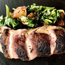 Aged Duck Breast - The image shows a black plate with sliced duck breast, featuring crispy, browned skin and a pink interior. Accompanying the duck are sautéed greens and small potatoes. The dish appears to be a prepared meal served in a restaurant setting, emphasizing the presentation of the meat and side vegetables.