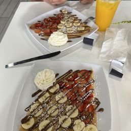 Orange Juice - Two plates of crepes topped with strawberries, bananas, chocolate drizzle, and powdered sugar, served with whipped cream. A person is using a smartphone, and a yellow drink is on the table.