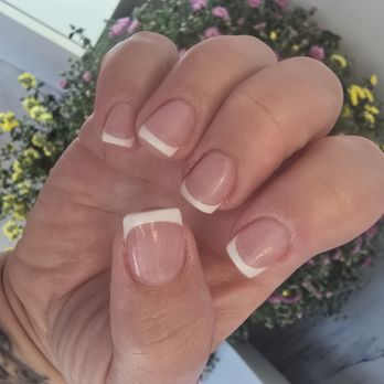 COCO Nail Bar - The image displays a close-up of a person's hand with nails painted in a French manicure style, characterized by natural pinkish nails with white tips. The background includes a blurred view of pink and yellow flowers, suggesting an outdoor or garden setting. The focus is on the well-maintained nails and the skin's texture. - Nail Salons Near Me - Algonquin, Illinois