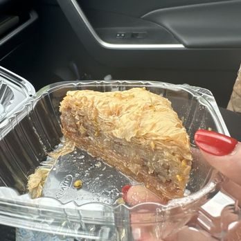 Cedar Cafe - Baklava - A slice of baklava, with visible layers of phyllo dough and chopped nuts, is contained in a clear plastic container. The container is held by a person with red-painted nails, seated inside a vehicle. The background shows part of the vehicle's interior, including the dashboard and door panel. - Greek Near Me - Norwell, Massachusetts