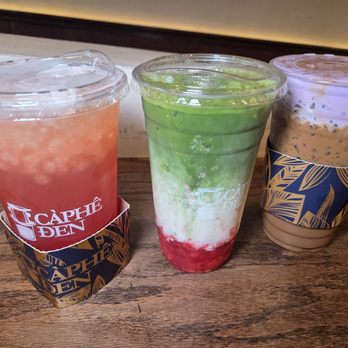Caphe Den - Three iced beverages in clear plastic cups with branded sleeves are arranged on a wooden table. The drinks include a pinkish-red drink, a layered green, white, and red drink, and a brown drink with a frothy top. Each cup has a plastic lid, and the drinks display different textures and colors, indicating a variety of flavors and ingredients. - Coffee & Tea Near Me - Flushing, New York
