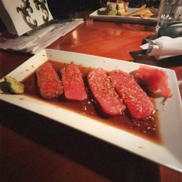 Tuna Crudo - The image shows a rectangular white plate with slices of raw fish, likely tuna, immersed in soy sauce, garnished with sesame seeds. A small portion of wasabi is placed on the side of the plate. The setting appears to be a restaurant with a dark, cozy ambiance, featuring a wooden table and additional dishes in the background.