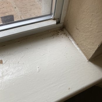 Texas Cleaning Services - Window sills were not cleaned - The image shows a close-up of a white-painted window sill with visible dust, dirt, and some debris. The window has a screen and a metal frame, with part of the exterior visible through the screen. The surrounding wall is textured and painted in a beige or light tan color. The window sill appears to be in need of cleaning. - Home Cleaning Near Me - Austin, Texas