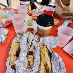 Super Migas Tacos - A table with foil-wrapped tacos, cups of water, coffee, and various sauces, set outdoors with a person in the background.