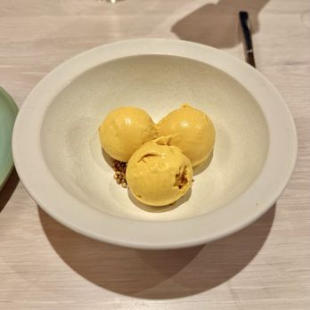 Fallow Kin - Pumpkin sesame ice cream - A white bowl contains three scoops of pumpkin sesame ice cream, which are yellowish in color. The ice cream is placed on a small bed of sesame seeds. The bowl is on a light-colored wooden table, with a black utensil visible in the background. The setting appears to be in a restaurant or cafe. - American Near Me - Cambridge, Massachusetts