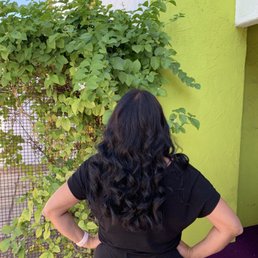 Long hair blow out with curls. - A woman with long, dark, curly hair stands outdoors with her back to the camera, next to a green wall and leafy plants.