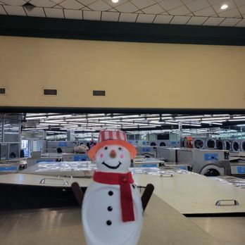 Laundry Palace - Snowman was here - A snowman-shaped figurine with a red scarf and striped hat is positioned on a table inside a laundromat, with washing machines visible in the background. - Laundry Services Near Me - Riverhead, New York
