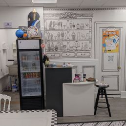 Checkin - The image shows the interior of a craft studio with a white ceiling and walls. A black fridge with a glass door is positioned next to a small counter, which has various small items on top. The wall behind features a detailed line drawing of a shelf filled with jars, bottles, and decorative objects, resembling a real shelf. To the right, there is a door with a poster attached, and a black stool with a stack of papers or magazines on it. The floor has a wood-like pattern, and part of a white table with a checkered edge is visible in the foreground.
