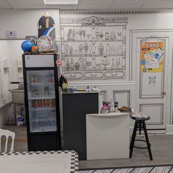 Crafts Zone Studio - Checkin - The image shows the interior of a craft studio with a white ceiling and walls. A black fridge with a glass door is positioned next to a small counter, which has various small items on top. The wall behind features a detailed line drawing of a shelf filled with jars, bottles, and decorative objects, resembling a real shelf. To the right, there is a door with a poster attached, and a black stool with a stack of papers or magazines on it. The floor has a wood-like pattern, and part of a white table with a checkered edge is visible in the foreground. - Paint & Sip Near Me - Brookline, Massachusetts