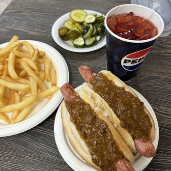The Lantern - Fries, pickles, two dogs all the way (mustard, onion, chili sauce) and a red birch beer. - Two hot dogs topped with chili sauce, French fries, a side of pickles, and a Pepsi soda on a dark wood table. - New American Near Me - Wanaque, New Jersey