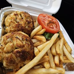 Jumbo Lump - The image shows a white takeout container with two crab cakes, French fries, a slice of tomato, and a small piece of lettuce. The crab cakes are golden brown with visible chunks of crab meat. The French fries are light golden and scattered around the crab cakes. The tomato slice is red with visible seeds, and the lettuce is green. The container is lined with foil at the bottom, and the lid is open, revealing the food inside.