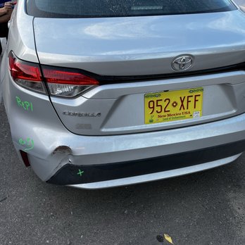 Caliber Collision - Damaged bumper - The image shows the rear view of a silver Toyota Corolla with visible damage to the lower left side of the bumper, which is dented and scratched. There are green handwritten markings on the left side of the bumper and near the taillight. The vehicle has a New Mexico license plate with the number 952 XFF. The car is parked on an asphalt surface with some fallen leaves nearby. The rear windshield appears dirty, and part of a person’s arm is visible on the left side of the image. - Body Shops Near Me - Rio Rancho, New Mexico