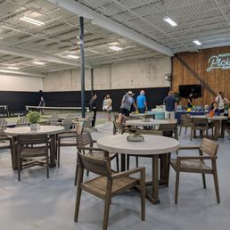 The image shows an indoor pickleball facility with multiple courts, some of which are in use by players. There is a seating area with round tables and wooden chairs in the foreground. A wooden wall with a neon sign reading 'Austin Pickle Ranch' is visible, along with a staircase leading to an upper level. The space has a high ceiling with exposed beams and bright lighting.