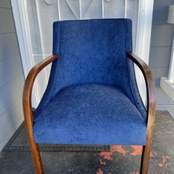 Sorto’s Furniture Repair & Caning Repair - A blue upholstered armchair with curved wooden armrests stands on a porch with a white door and decorative metalwork in the b...