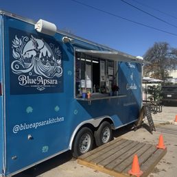A blue mobile food truck with the Blue Apsara logo and branding for Cambodian cuisine is parked in an outdoor lot. The truck has a serving window with a small menu display and potted plants nearby. Orange cones and a wooden platform are positioned in front of the truck, with parked cars and trees visible in the background.