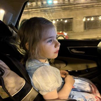 Broad Brook Opera House - A young girl with light brown hair, styled in loose waves, is seated in a car seat inside a vehicle. She is wearing a dress with puffy sleeves and a shiny, patterned skirt. Her gaze is directed out the window, showing a thoughtful or curious expression. The background outside the window features city lights, a bridge, and a passing red vehicle, indicating an urban setting at night. - Venues & Event Spaces Near Me - Broad Brook, Connecticut