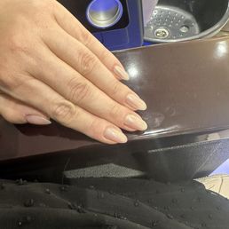 Glazed donut nails - The image shows a person's left hand with well-maintained, natural-colored nails resting on a dark brown surface. In the background, there is a computer monitor with a blue and black design and a sink with a metallic drain, indicating an indoor setting. The person's clothing includes a black fabric with small textured details.