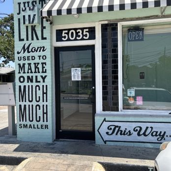 Tiny Pies - The image shows the exterior of a small business with a mural on the left side that reads, "JUST LIKE Mom USED TO MAKE ONLY MUCH MUCH SMALLER." The mural is painted on a light-colored brick wall. To the right of the mural, there is a glass door with the number 5035 above it, and a sign on the door. An arrow painted on the wall points to the right with the words "This Way." A black-and-white striped awning extends over the entrance, and a small sign indicating the business is open hangs in the window. The sidewalk in front includes a small ramp and parking space, with part of a white vehicle visible on the right. - Desserts Near Me - Austin, Texas