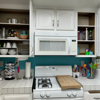 Clearing Space - After - A kitchen with white cabinetry, including upper cabinets and open shelves holding dishes, cups, and kitchen items. A microwave is mounted above a white stove with a wooden cutting board on top. The counter has a paper towel holder, a spice rack, and containers with utensils and colorful straws. The background wall is painted teal, and there are electrical outlets visible on the backsplash. - Home Organization Near Me