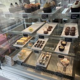 The image shows a glass display case filled with an assortment of baked goods such as cupcakes, cookies, and slices of cake. The items are arranged on white trays with small handwritten labels indicating their names and prices. The case is situated in a bakery or cafe setting, with a reflection of the outside visible through the glass. The baked goods include decorated cupcakes, cookies, and other sweet treats, some with toppings or frosting.