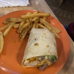 Buffalo Chicken Mac & Cheese Sandwich - A burrito with a flour tortilla wrap, filled with ingredients such as chicken, lettuce, cheese, and possibly other vegetables, is placed on an orange plate. Accompanying the burrito are French fries, some of which are slightly browned. The plate is on a wooden table, with utensils wrapped in a napkin visible in the background, indicating a casual restaurant environment.