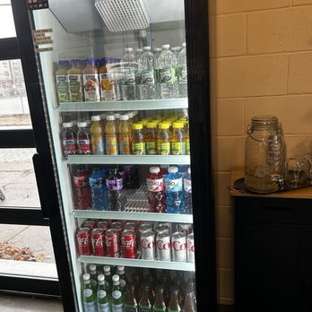 LIFE Cafe - Cold Drinks - A glass-front refrigerator contains multiple shelves filled with bottled beverages such as water, juice, soda, and energy drinks. The fridge is located indoors, next to a brick wall and a counter with a jar and glassware. The setting appears to be a cafe or casual dining area, with natural light coming from a nearby window. - Coffee & Tea Near Me - Pawtucket, Rhode Island