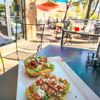 Daily Jam - Breakfast & Brunch Restaurant - Two pieces of avocado toast topped with diced tomatoes, crumbled cheese, and herbs are placed on a white rectangular plate. The setting is an outdoor cafe with tables, chairs, and colorful umbrellas visible in the background, along with trees and sunlight. - New American Near Me - Tempe, Arizona