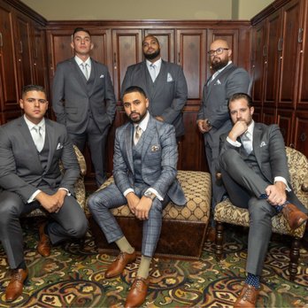 Savage Suiting - All suits from Savage! Quality. - Seven men dressed in suits are positioned in a wood-paneled room with patterned carpet, some seated and others standing, in a formal group photo. - Formal Wear Near Me - Placentia, California