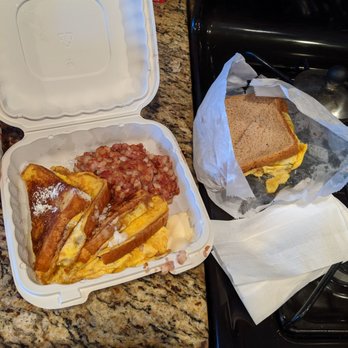King's Cafe - Breakfast! - A foam takeout container on a granite kitchen countertop contains French toast, scrambled eggs, and bacon. The French toast appears golden brown with powdered sugar, and the eggs are partially visible. Next to the container, a sandwich with bread and eggs is wrapped in paper, resting on a napkin. The scene suggests a breakfast meal prepared for takeout or casual dining at home. - Cafes Near Me - Washington, DC, District of Columbia