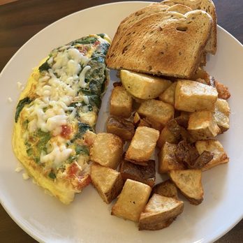 Lemon Leaf Cafe - Veggie omelet - A breakfast plate includes a cheese and vegetable omelet with visible spinach, tomatoes, and melted cheese, served alongside toasted slices of bread and roasted potato cubes. The plate is on a dark wooden table, with part of a person's arm and shirt visible in the background. - Breakfast & Brunch Near Me - Largo, Florida