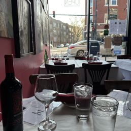A restaurant interior with tables set for dining, including glasses, bottles, and menus, with a large window revealing parked cars and buildings outside.