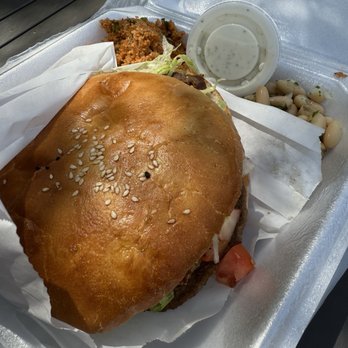 Sultan Kebab - A foam takeout container contains a burger with a sesame seed bun, lettuce, tomato, and other fillings. Alongside, there is fried chicken, white beans, and a small cup of creamy dipping sauce. The meal is placed on a dark surface, likely outdoors or on a picnic table. - Turkish Near Me - Charlottesville, Virginia