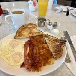 American Breakfast - A breakfast plate includes two fried eggs, crispy bacon, hash browns, and toasted bread slices, with coffee and juice on the table.