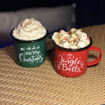 Edge Rooftop - Two ceramic mugs with holiday designs are placed on a woven table surface. The green mug features 'Merry Christmas' with a Santa sleigh graphic, and the red mug displays 'Jingle Bells' with a gift and bell illustration. Both mugs are filled with hot chocolate, topped with whipped cream, and garnished with marshmallows and festive sprinkles. - Lounges Near Me - Austin, Texas