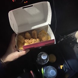 App Sampler - A hand is holding an open box of fried chicken inside a vehicle. The car's dashboard and climate controls are visible in the background, along with two water bottles placed in the cup holders. The scene appears to be at night, with the car's interior lights illuminating the area.