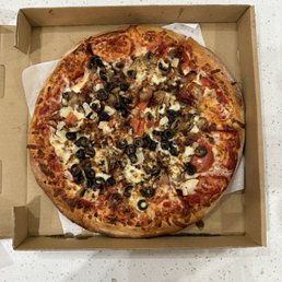 Deluxe Pizza - A round pizza with a golden-brown crust is placed inside a cardboard pizza box on a white speckled countertop. The pizza is topped with black olives, sliced mushrooms, chopped onions, and pepperoni slices, with melted cheese covering the toppings. The pizza appears freshly baked and ready for pickup or delivery.