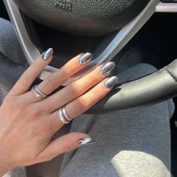 De Paris Nails - Chrome dip w tip - A hand with metallic silver painted nails and multiple rings is placed on a car steering wheel, with the person's lap visible in the background. - Nail Salons Near Me - Centerville, Massachusetts
