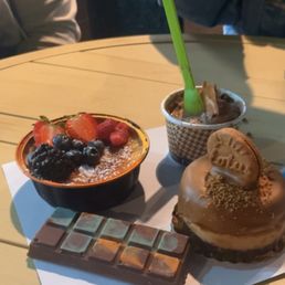 Crème brûlée, Biscoff, cheesecake, Dubai, chocolate, chocolate ice cream - A selection of desserts on a white paper sheet includes a chocolate bar, a fruit tart with strawberries and blueberries, a chocolate-covered pastry with a cookie, and a cup of ice cream with a green spoon.