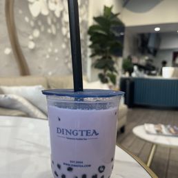 Taro Milk Tea - A plastic cup of taro milk tea with black tapioca pearls at the bottom, branded with Ding Tea, placed on a white table in a modern cafe setting.
