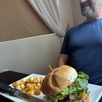 Stem & Stone - Burger - A man with glasses, a beard, and gray hair is smiling while sitting at a table. In front of him is a white plate with a large burger containing lettuce, tomato, cheese, and meat, along with a side of fried potato cubes. The background includes a beige wall and a window with white curtains. - New American Near Me - Bastrop, Texas