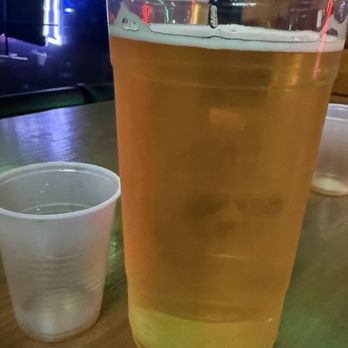 Northside Tap - A large clear plastic cup filled with light-colored beer, placed on a wooden table in a dimly lit setting with colorful lights in the background. - Bars Near Me - Valparaiso, Indiana