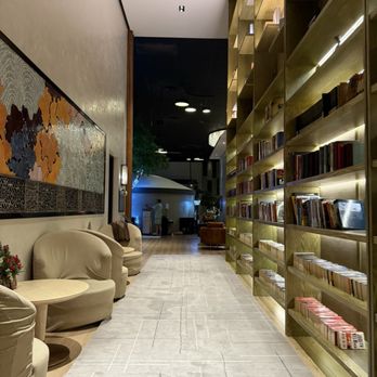 Go Place - The image depicts a lounge area featuring beige, plush armchairs and a small round side table. A large wooden bookshelf with multiple compartments filled with books lines the right side of the space. The wall on the left has a decorative artwork with a map design, and the floor is covered with a light-colored rug. In the background, there are additional seating areas and a dark ceiling with circular lighting fixtures, suggesting a comfortable, inviting environment. - Day Spas Near Me - Markham, Ontario