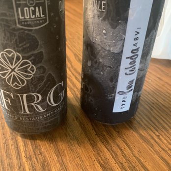 The Local - Another look at the drinks - Two black cans are placed on a wooden table with visible wood grain. The can on the left has a logo with the words 'LOCAL' and 'BAYONNY,' along with a clover design. The can on the right has a white label with handwritten-style text that reads 'TYPE Pecan Tisoda ABV.' Both cans have condensation on their surfaces. - Cocktail Bars Near Me - Babylon, New York