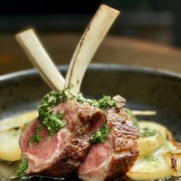 Lamb Chops - The image shows two lamb chops with a pink interior and seared exterior, garnished with chopped herbs. The lamb is placed on a bed of sliced potatoes, and the dish is served in a dark bowl. The focus is on the meat and garnishes, with a blurred background.