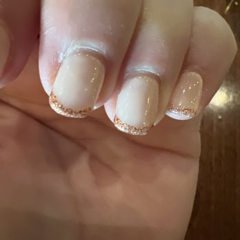 4J Nails & Spa - Builder gel wi/nude color & orange sparkle French. last time in was a month ago, NONE were broken or chipped. Builder gel is the bomb. - The image shows a close-up of a hand with neatly manicured nails. The nails are painted with a light nude polish and have a gold glitter French tip design. The background includes a wooden surface, and the lighting highlights the glossy finish of the nails. - Waxing Near Me - Albuquerque, New Mexico