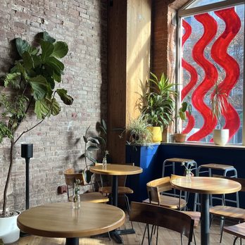 Le Petit Monstre - Ambiance - A cafe interior features wooden tables and chairs, potted plants on the tables and window sill, and a large window with red decorative glass panels. - Patisserie/Cake Shop Near Me - Brooklyn, New York